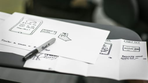 Three sheets of paper with hand-drawn sketches and notes, including diagrams of mobile interfaces, lie on a dark desk beside a black Sharpie marker.